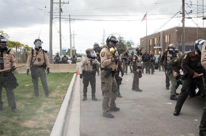 An Illinois State Police trooper, center, points a pepper ball gun at photojournalist Jon Stegenga. The officer shot him in the knee and thigh with the chemical irritant munitions amid protests outside a federal facility in Broadview on Nov. 1, 2025.
