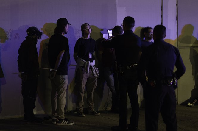 Journalist Melanie Buer, in middle wearing her press credential, stands facing police as an officer takes her photo. Buer and other press were detained by Los Angeles Police Department officers while covering a protest in LA on Aug. 8, 2025.