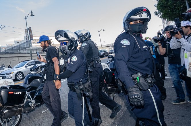Reporter Lexis-Olivier Ray, at left, is detained and cited by police for stepping into the street while documenting a workers’ rights and immigration protest in Los Angeles, California, on May 1, 2026.