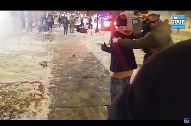 Status Coup reporter Tina-Desiree Berg, in hat at center right, as she is grabbed from behind and thrown to the ground by a federal officer while reporting on a protest outside a hotel in Minneapolis, Minnesota, on Jan. 25, 2026.