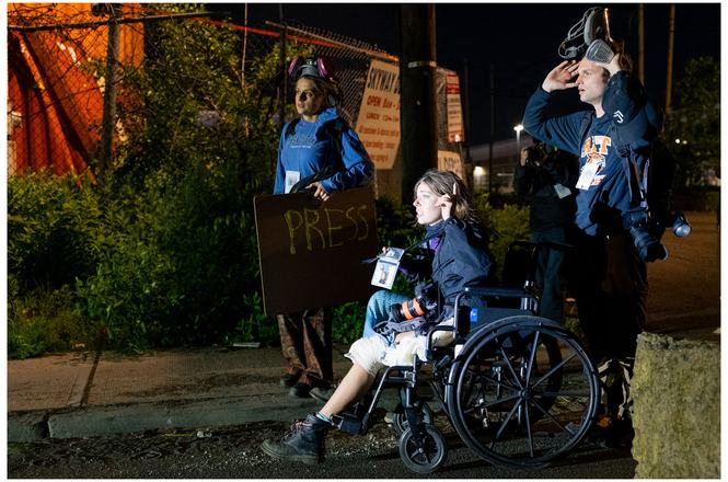 Photojournalist Angelina Katsanis, in wheelchair, negotiates to exit through police lines after being injured at a May 30, 2026, immigration protest in Newark, New Jersey. Her gear bag was stolen that night, and an officer was later charged with theft.